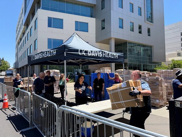Team members standing near a UC Davis Health tent unloading and moving boxes of medical supplies from pallets and a truck in an outdoor staging area at the North Addition Office Building